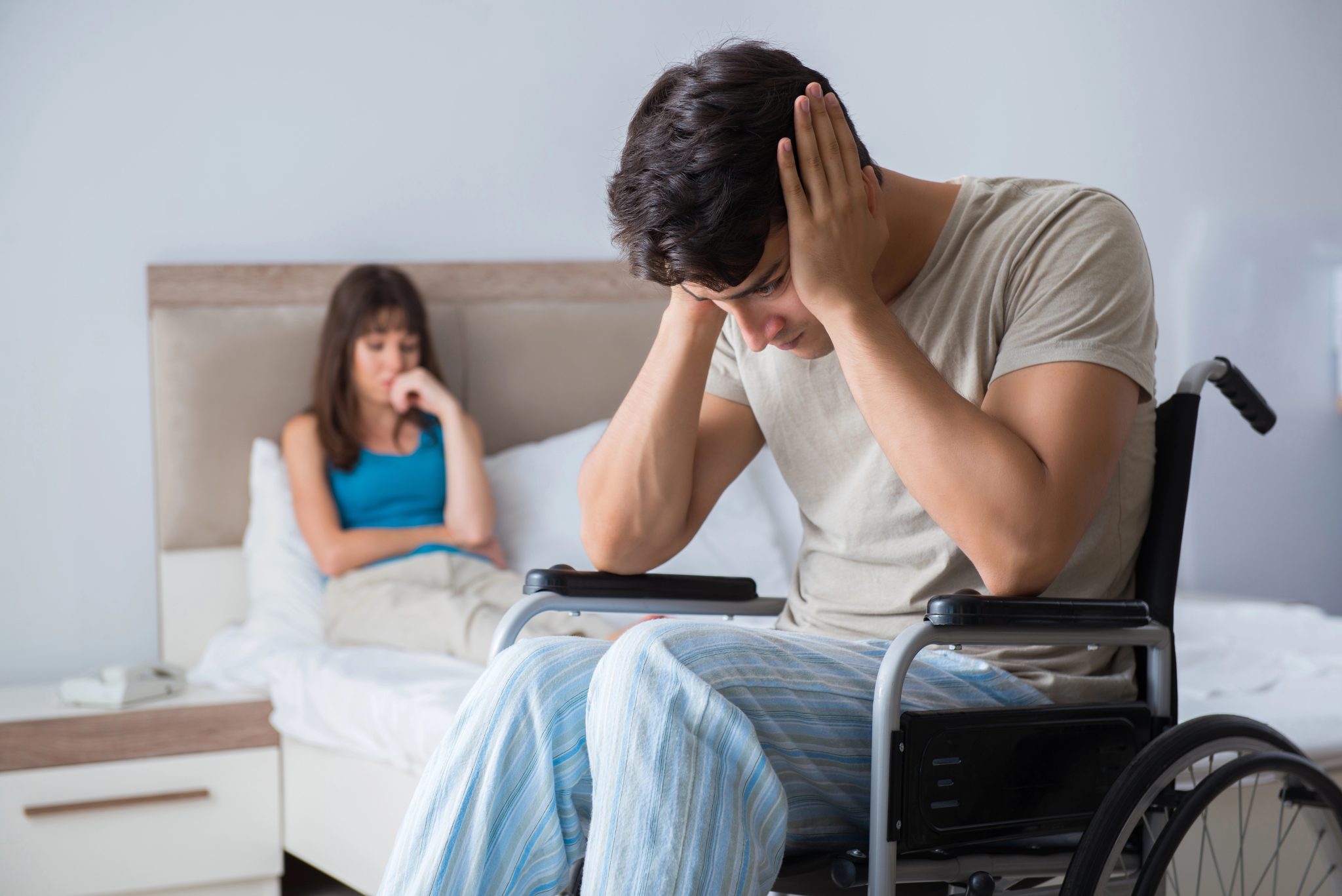 Man in a wheelchair holds his head in distress in a bedroom, while a woman sits on the bed in the background looking worried. 3 effects of crush injury on your life in Sartell | Gabrielson Law Offices, LTD 3 effects of crush injury on your life in Sartell | Gabrielson Law Offices, LTD